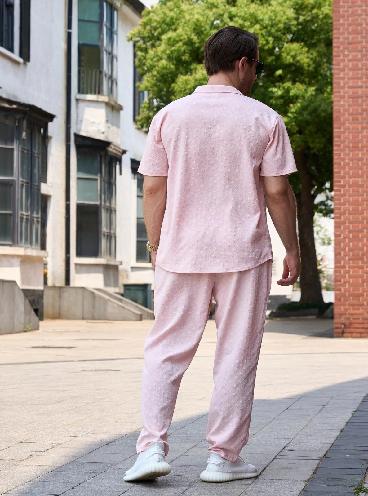 Person wearing a pink outfit walking on a sidewalk with buildings and trees in the background