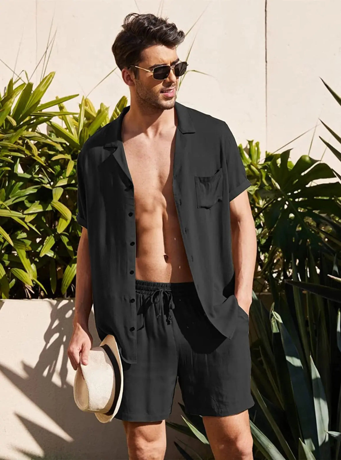 Man wearing a black shirt over black shorts with sunglasses and a hat, standing outdoors.
