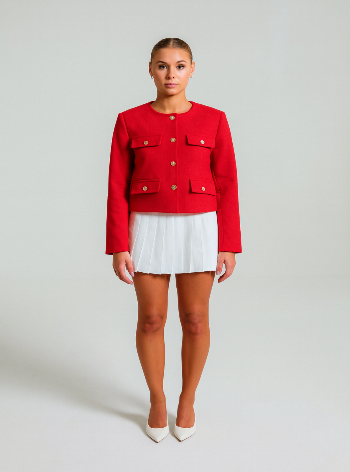 Model in a sleek ponytail wearing a red textured Jacket with gold buttons, a white pleated skirt and white pointed heels standing in front of a white background.