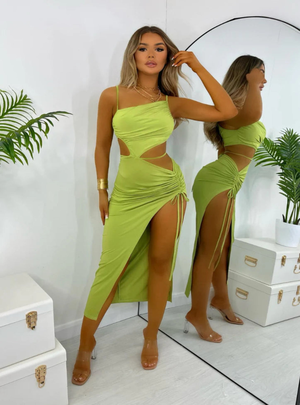 Woman in a bright green dress standing in front of a mirror, with plants and white boxes in the background.