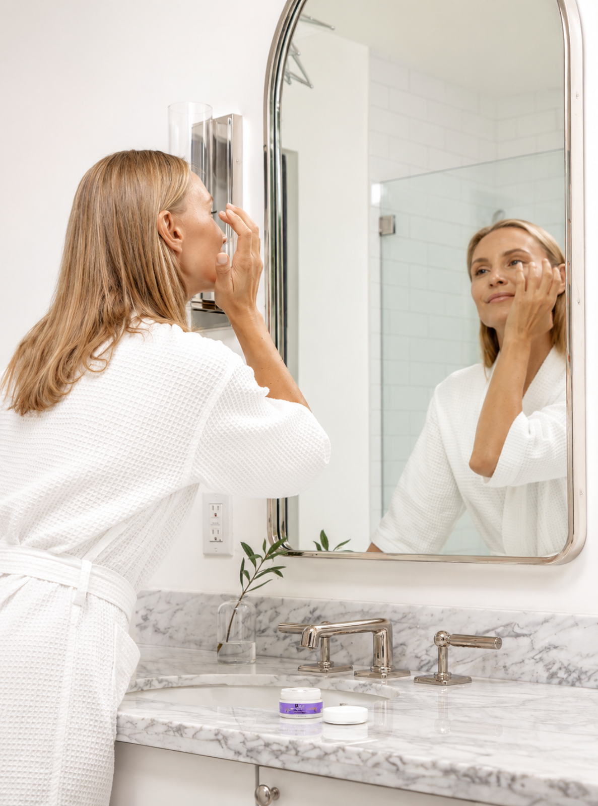 A woman in a white robe applies skincare cream to her face while looking at her reflection in a bathroom mirror above a marble countertop.