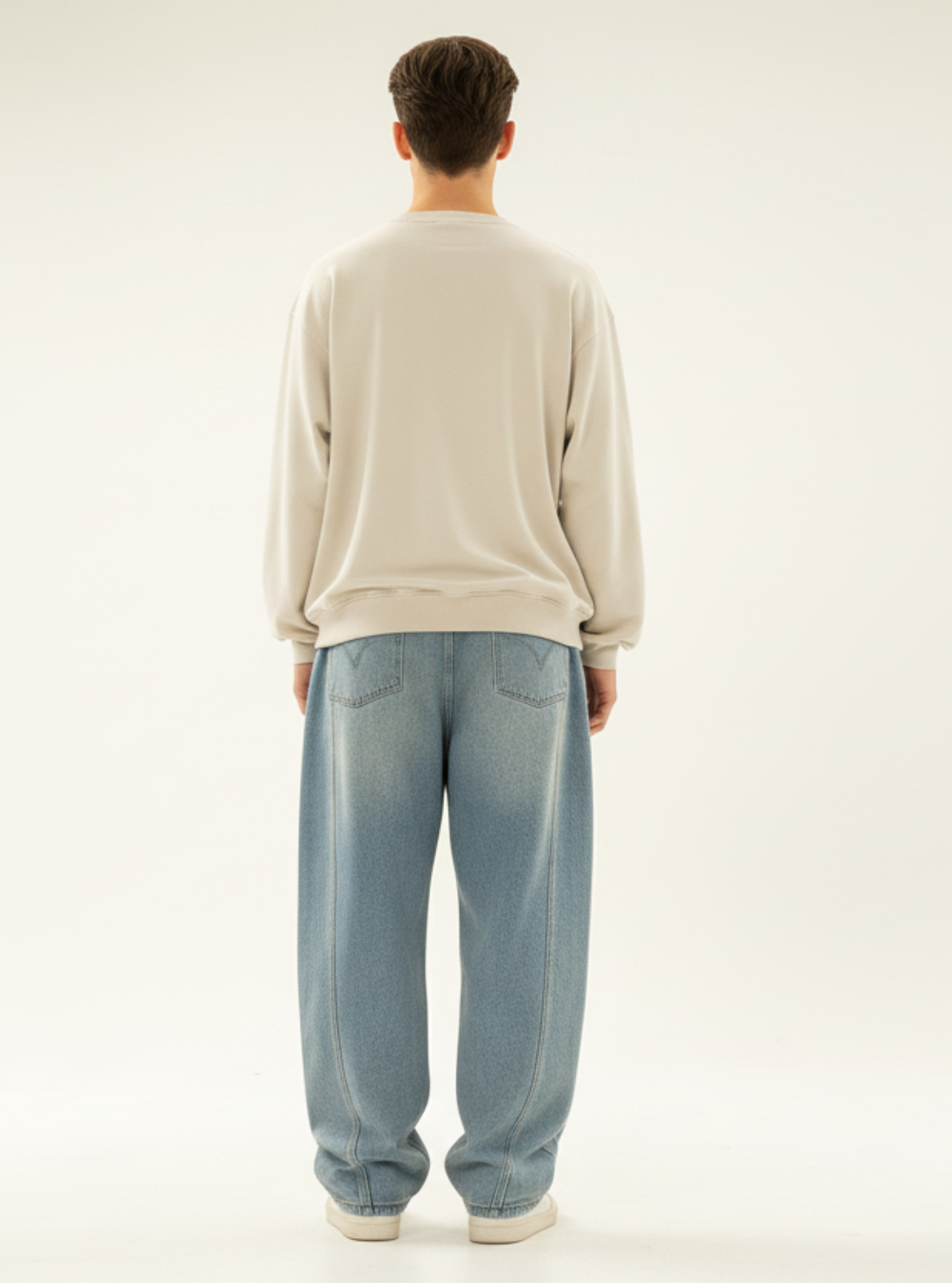 Person wearing a beige sweater and blue jeans standing against a plain background