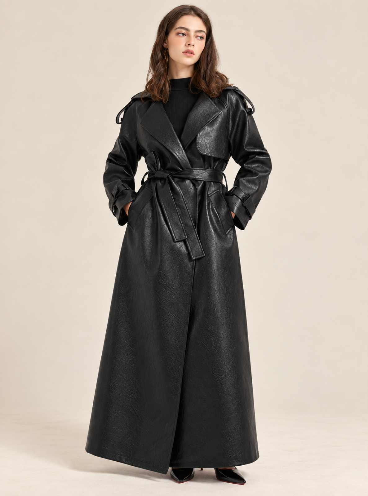A woman modeling a long black faux-leather trench coat with a matching waist tie belt, styled over a black top, against a plain beige studio background.