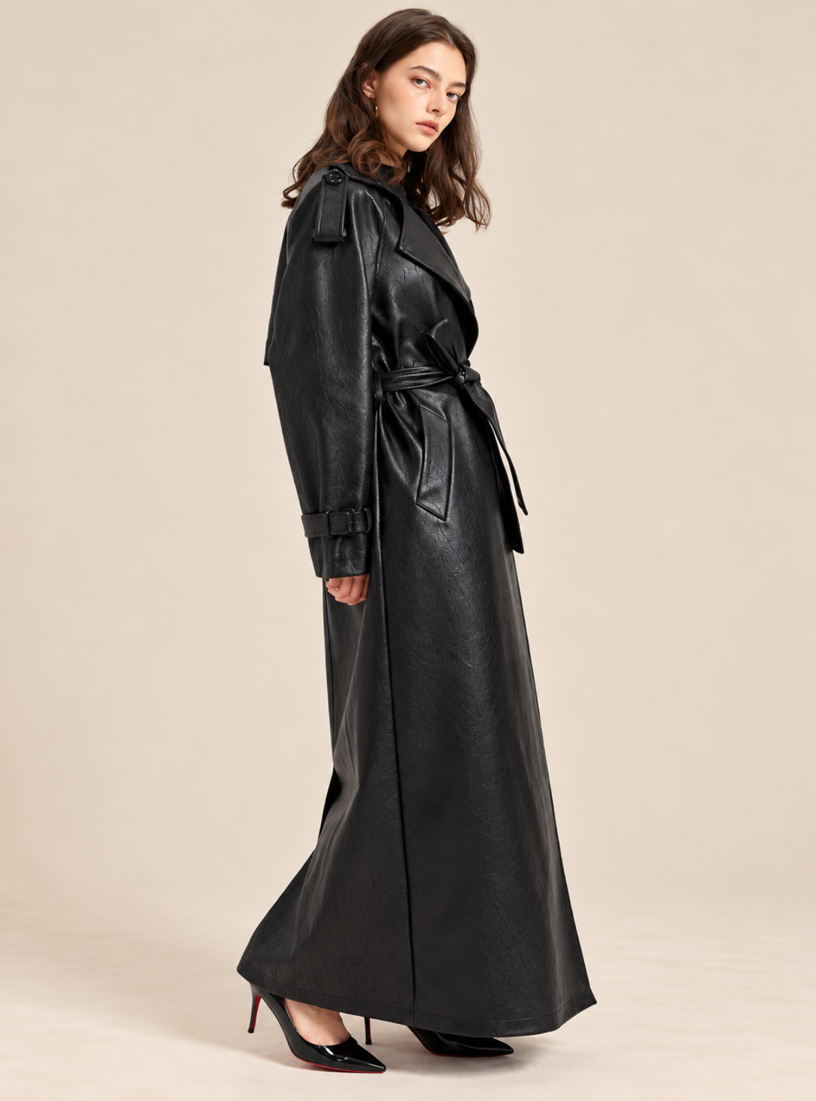 A woman modeling a long black faux-leather trench coat with a tie waist and shoulder epaulets, paired with black pointed-toe heels, against a solid beige studio background.