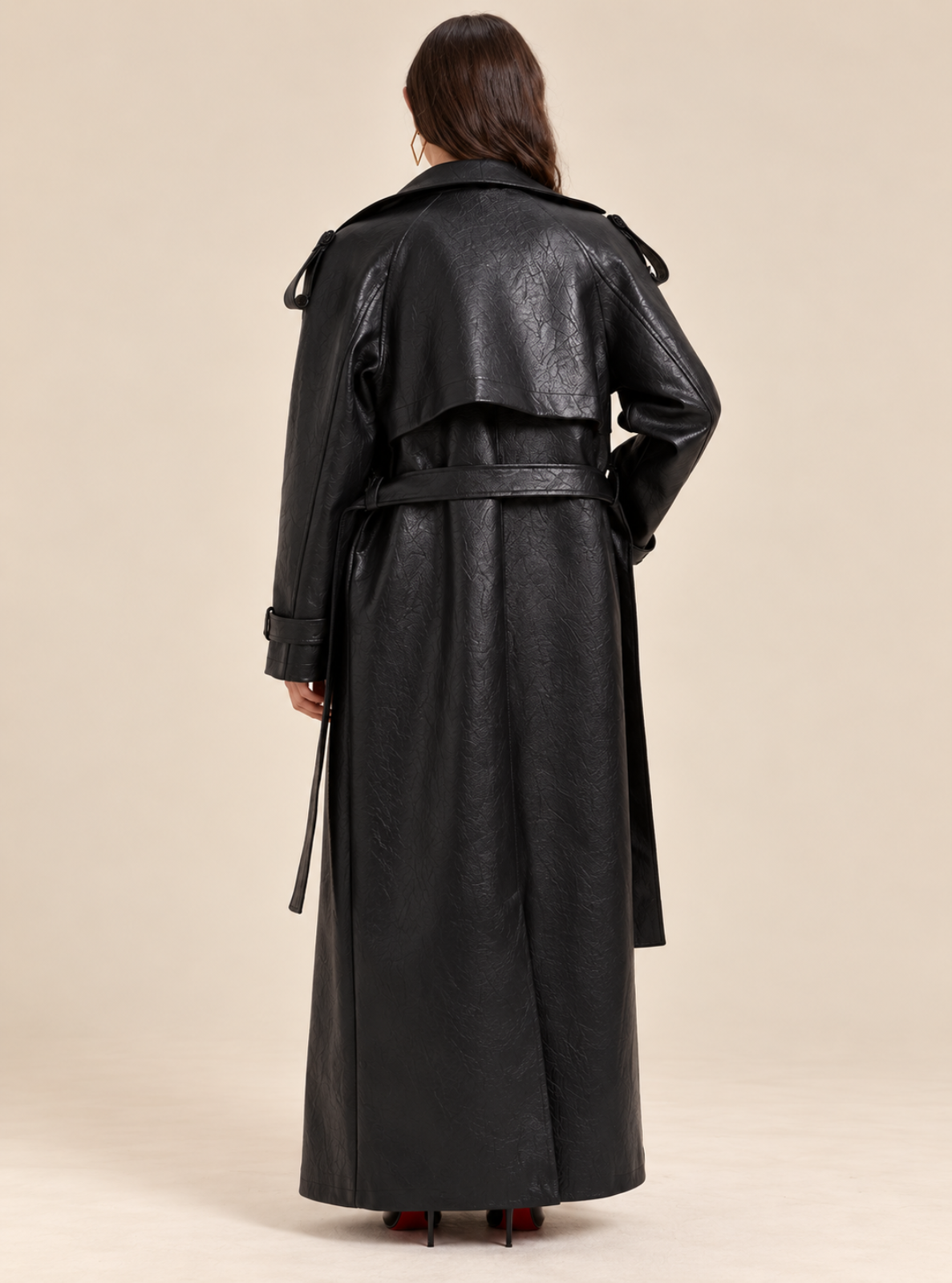 A model viewed from the back wearing a long black faux-leather trench coat with a matching waist belt, set against a solid beige studio background.