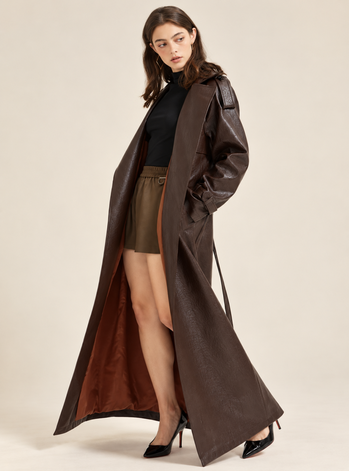A model wears a long, dark brown faux-leather trench coat with a contrasting brown satin lining, styled over a black top, olive green mini skirt, and black patent heels against a solid beige studio background.