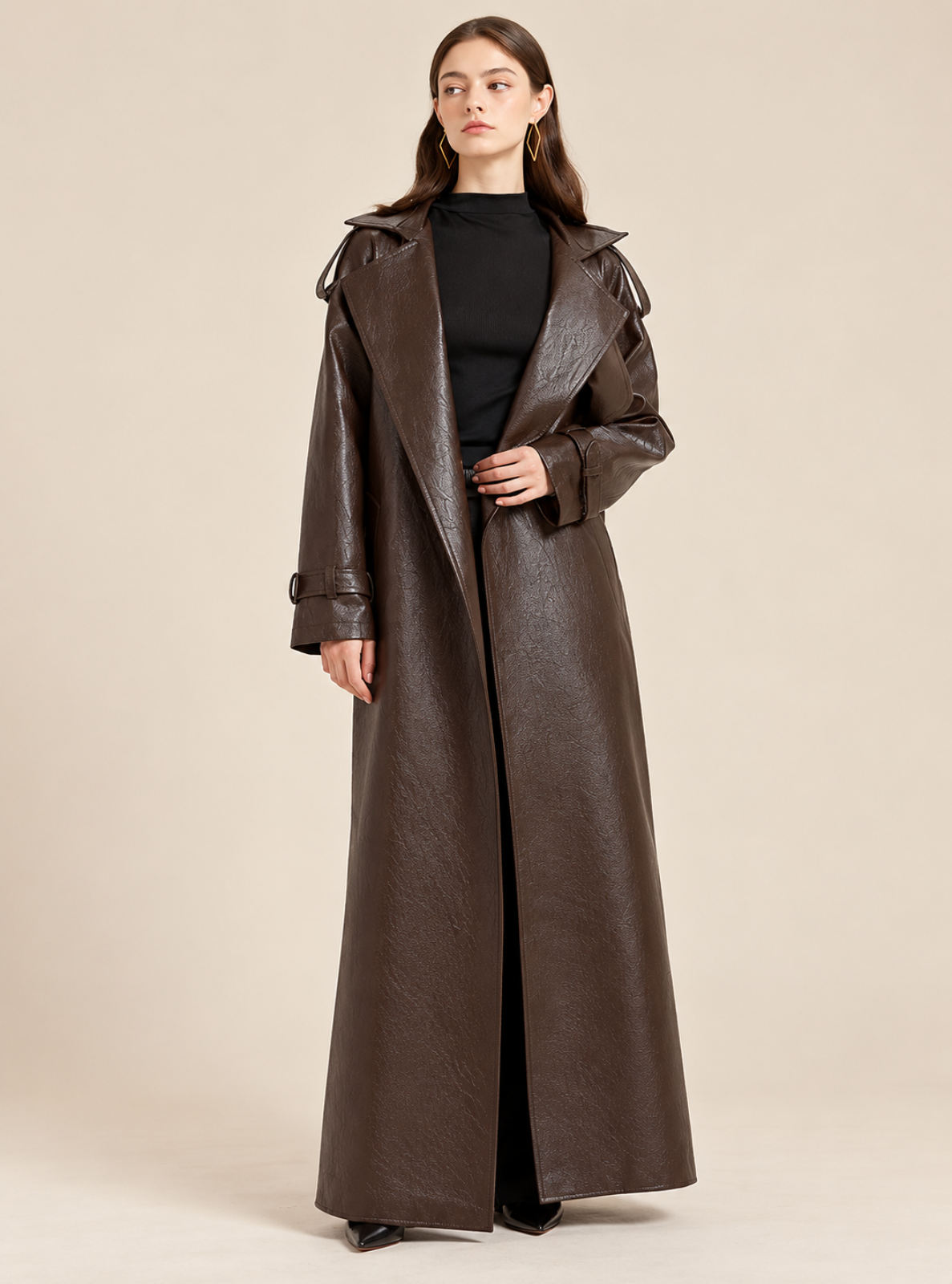 A woman modeling a long, dark brown faux-leather trench coat over a black top and black pants, standing against a solid beige studio background.