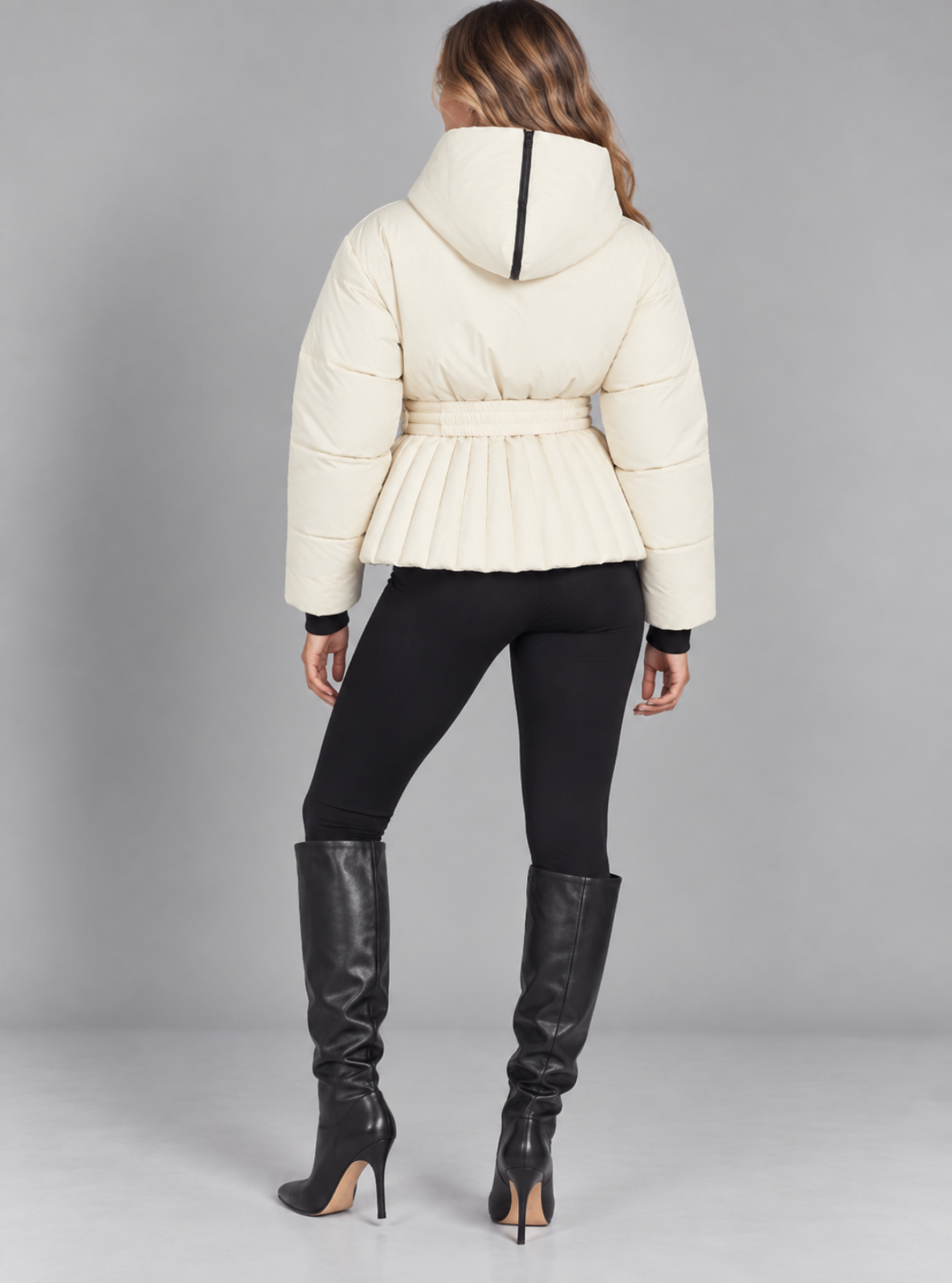 A woman shown from the back wearing a cream quilted puffer jacket with a hood and waist tie, black leggings, and black knee-high heeled boots against a light gray studio background.