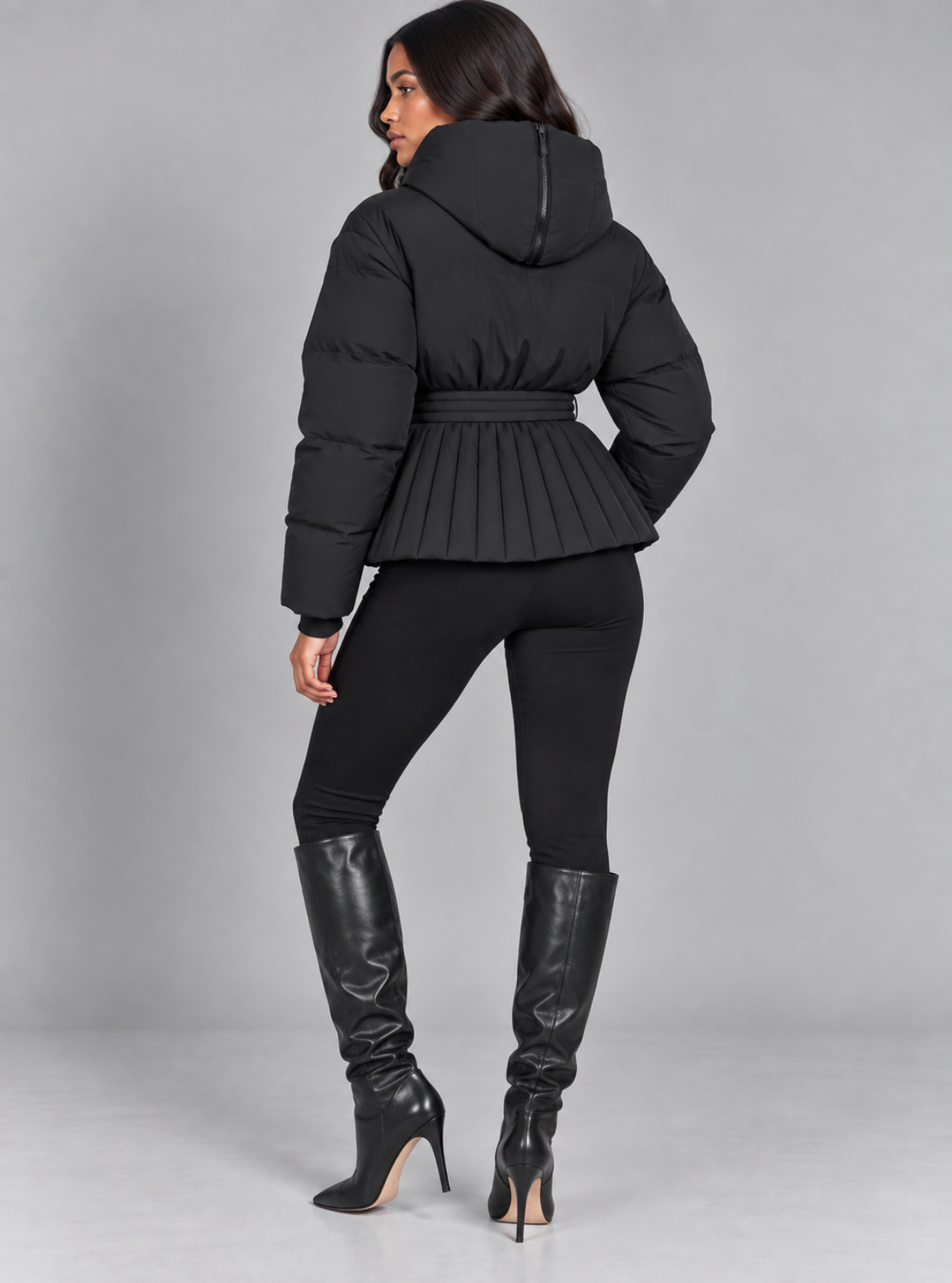 A woman shown from the back wearing a black hooded puffer jacket with a pleated peplum waist, black leggings, and black knee-high heeled boots against a light gray studio background.