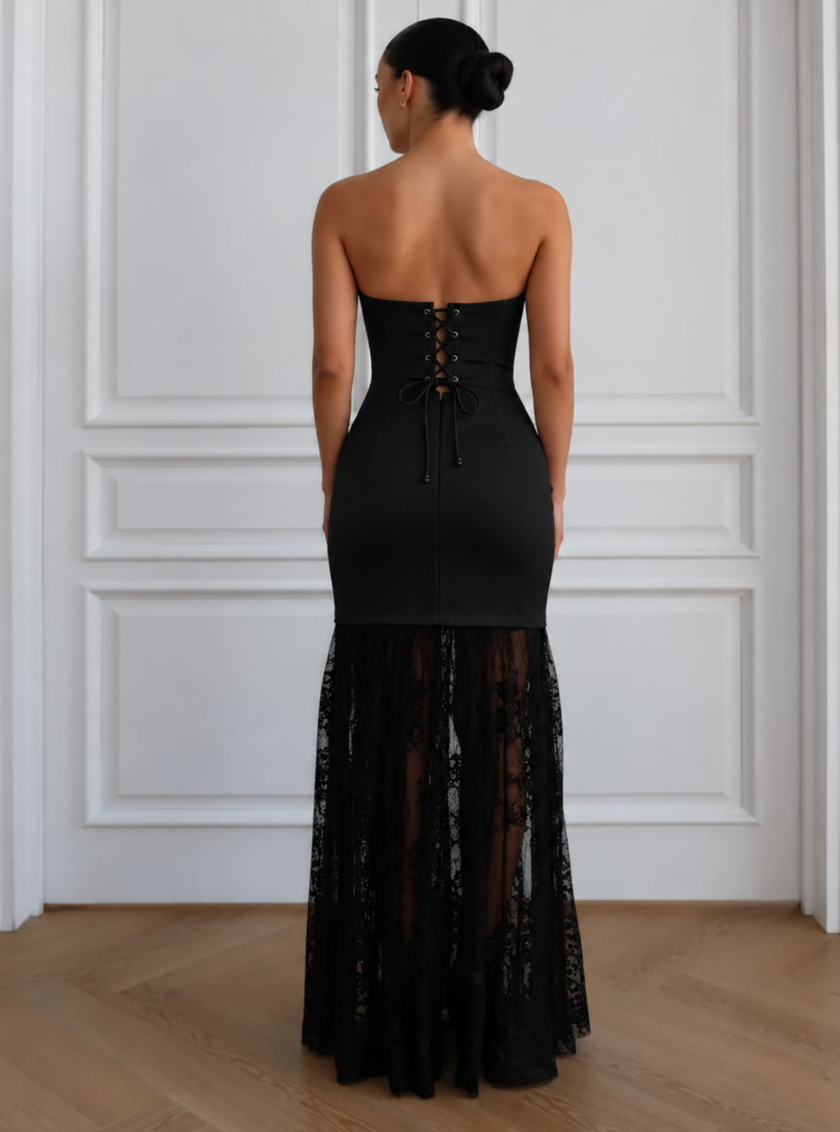 A woman viewed from the back wearing a strapless black gown with a fitted corset-style back, lace-up detailing, and a sheer lace mermaid skirt, standing in front of a white paneled wall.