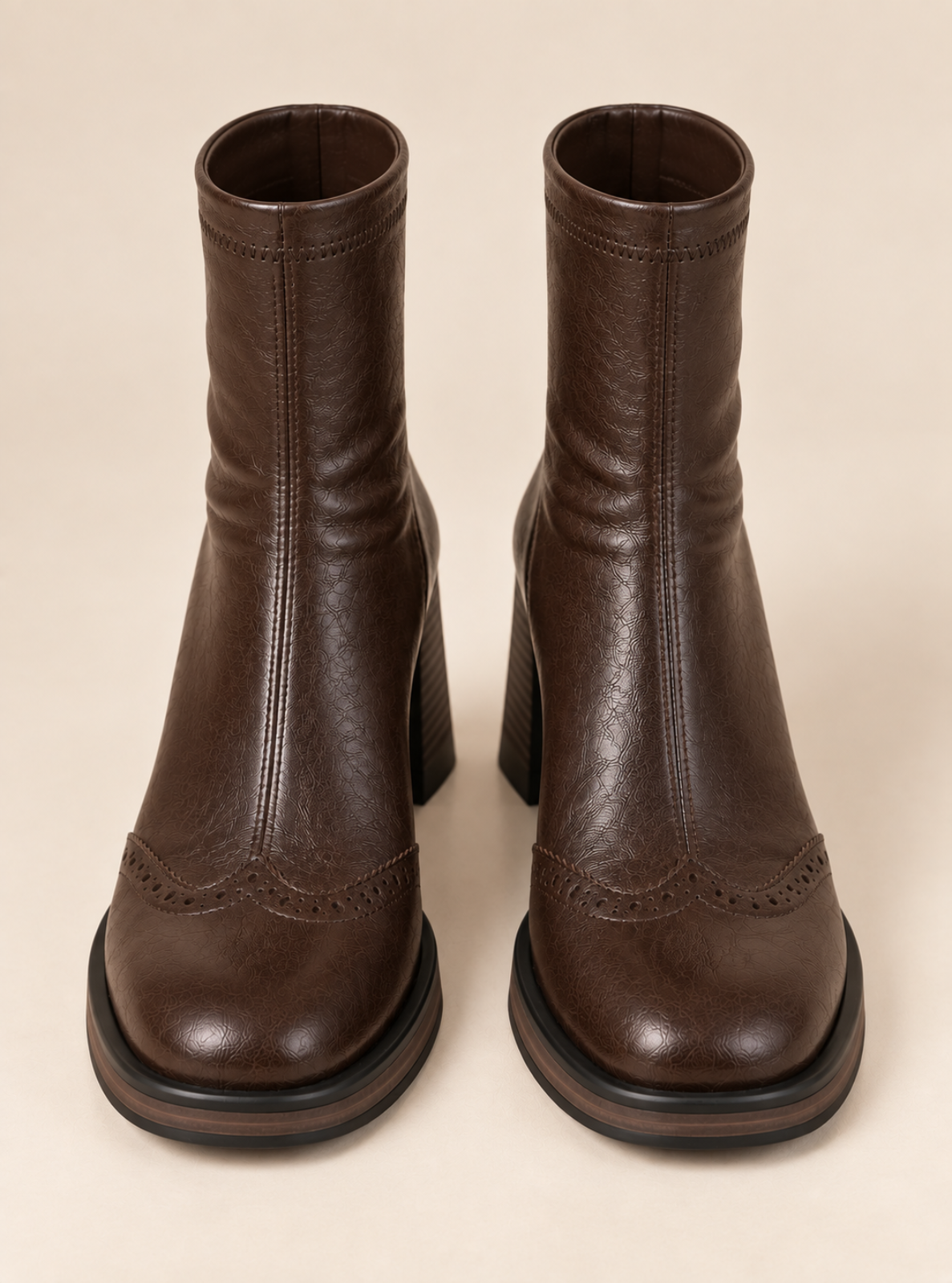 A pair of dark brown leather ankle boots with block heels and decorative brogue detailing on the toes, shown against a solid beige background.