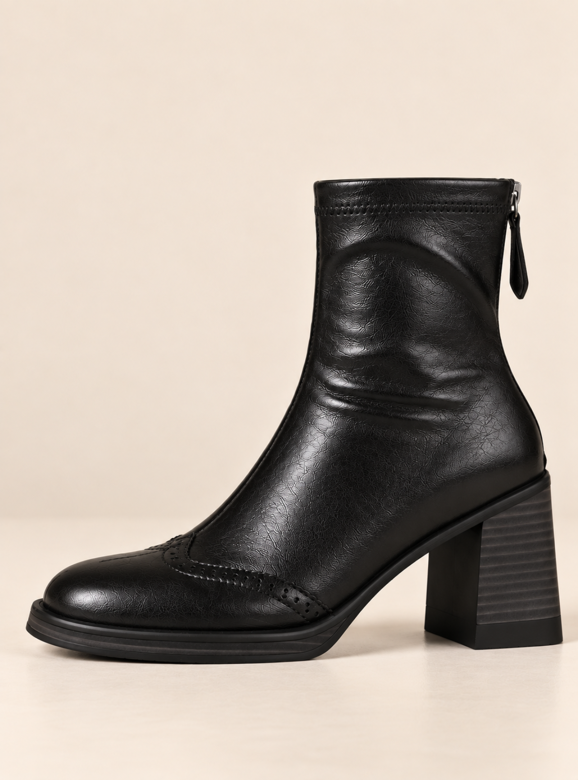 A black leather ankle boot with a chunky block heel, brogue detailing on the toe, and a side zipper, shown against a solid beige background.