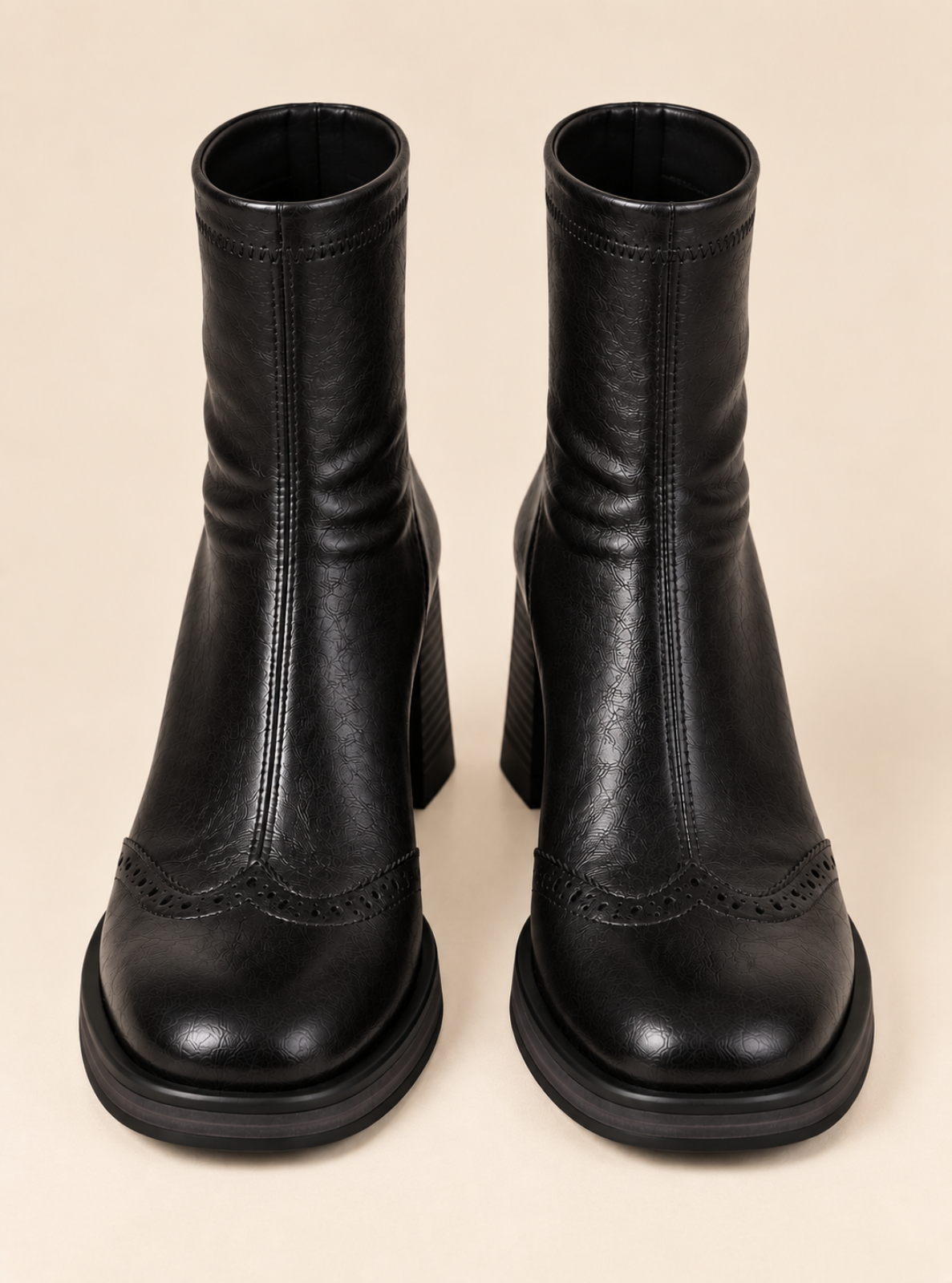 A pair of black leather ankle boots with a block heel and decorative brogue detailing on the toe, shown from a front-facing perspective on a solid beige background.