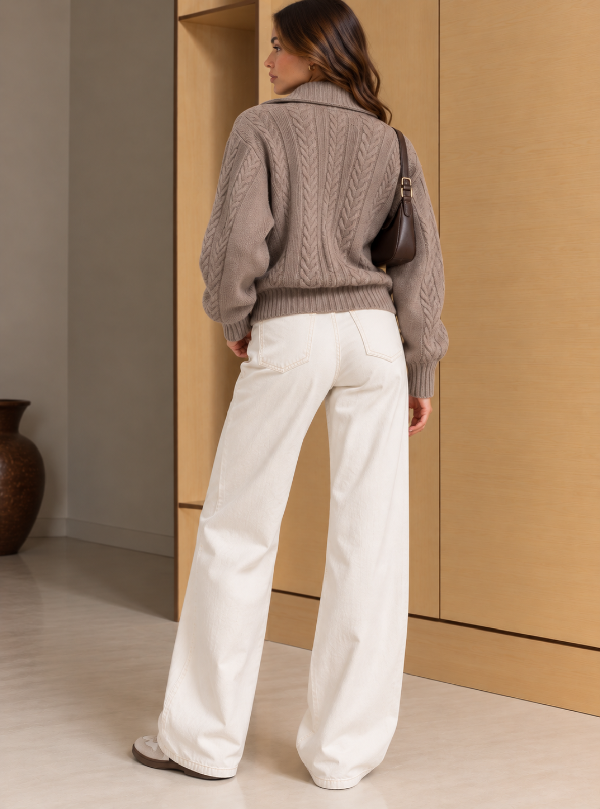 A woman shown from the back wearing a taupe cable-knit sweater, white wide-leg trousers, and carrying a dark brown shoulder bag in an indoor setting with beige walls.
