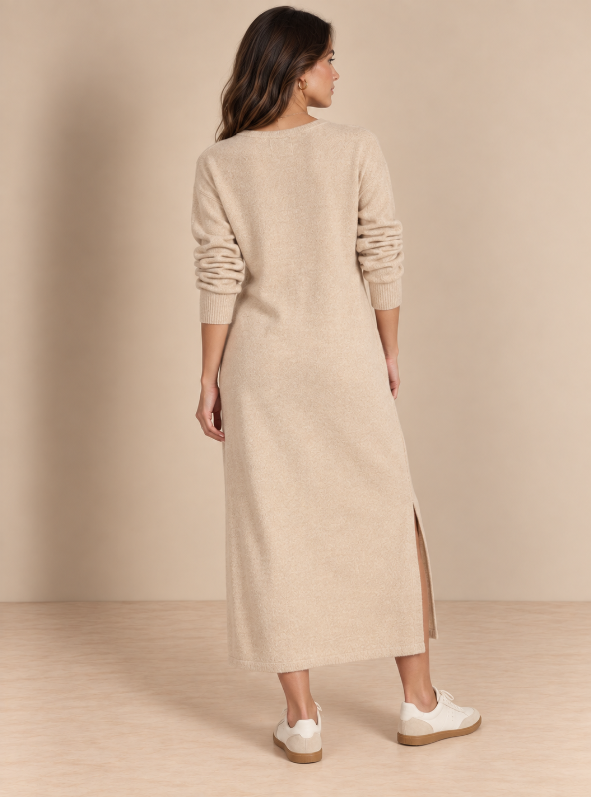 A woman is shown from the back wearing a long-sleeve, knee-length beige knit sweater dress with side slits, paired with white sneakers, against a neutral beige studio backdrop.