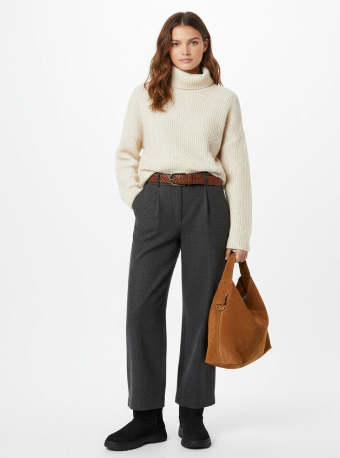 Woman wearing a cream sweater, gray pants, and holding a brown bag on a white background