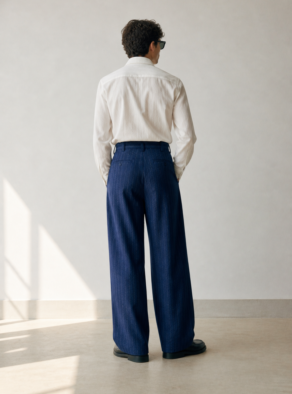 Person wearing a white shirt and blue pants standing in a room with a light gray wall and wooden floor.