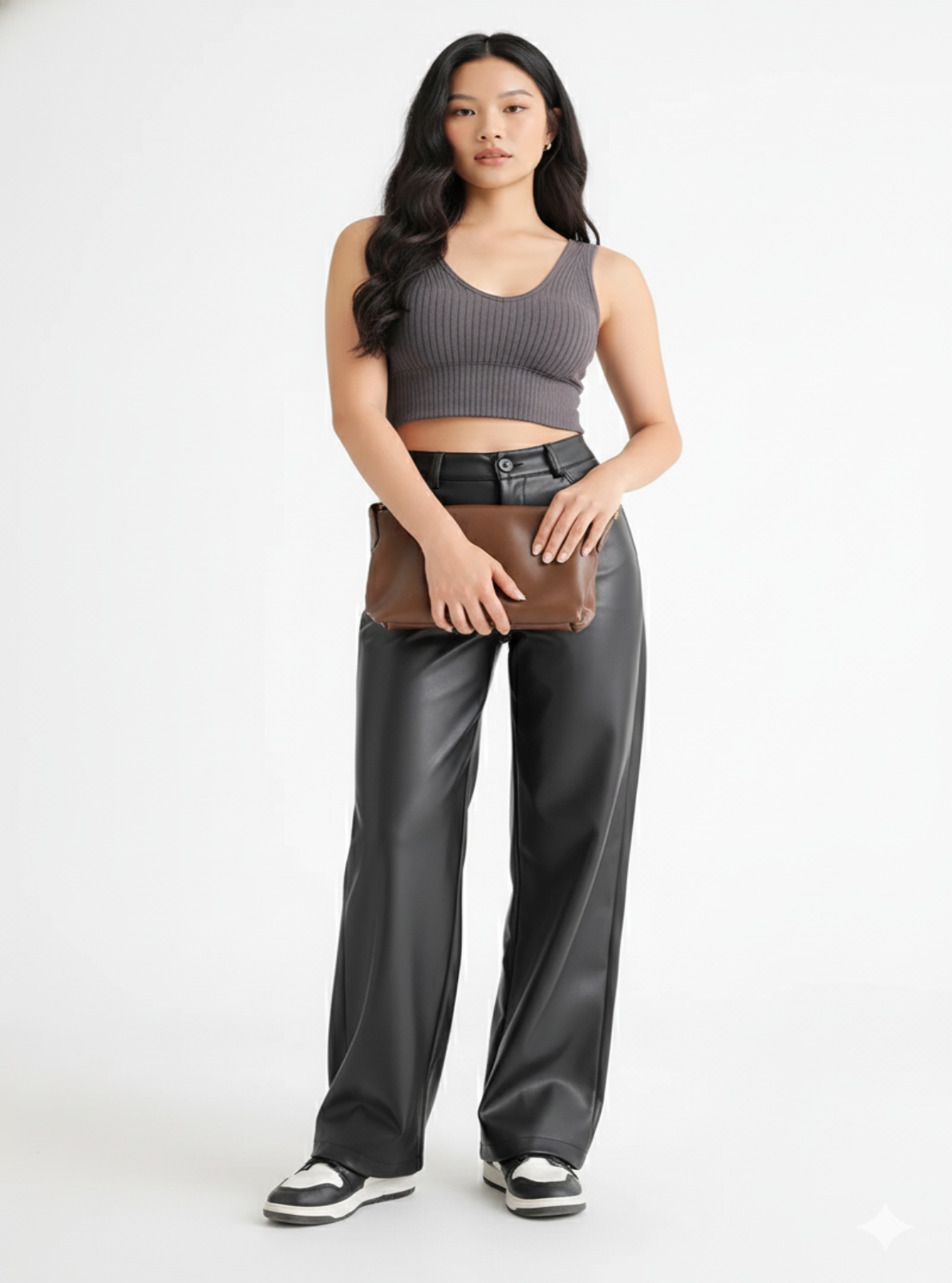 Woman wearing a gray tank top and black pants holding a brown leather jacket on a white background