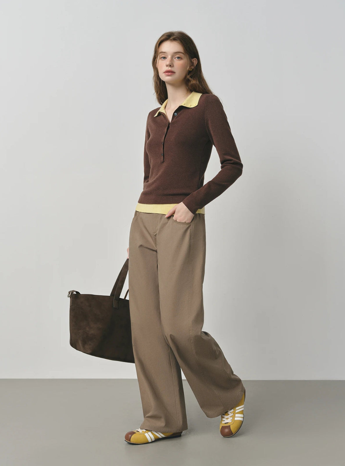 Woman wearing a brown outfit with a yellow shirt, holding a brown bag against a gray background