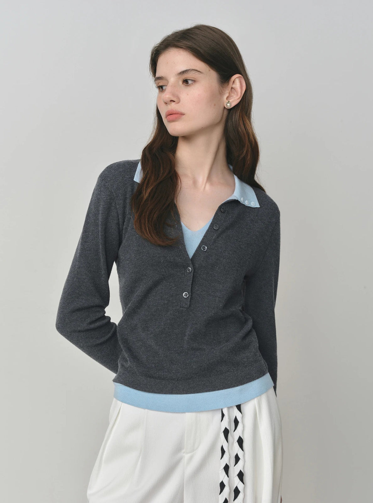 Woman wearing a gray cardigan over a light blue shirt with a white skirt on a plain background