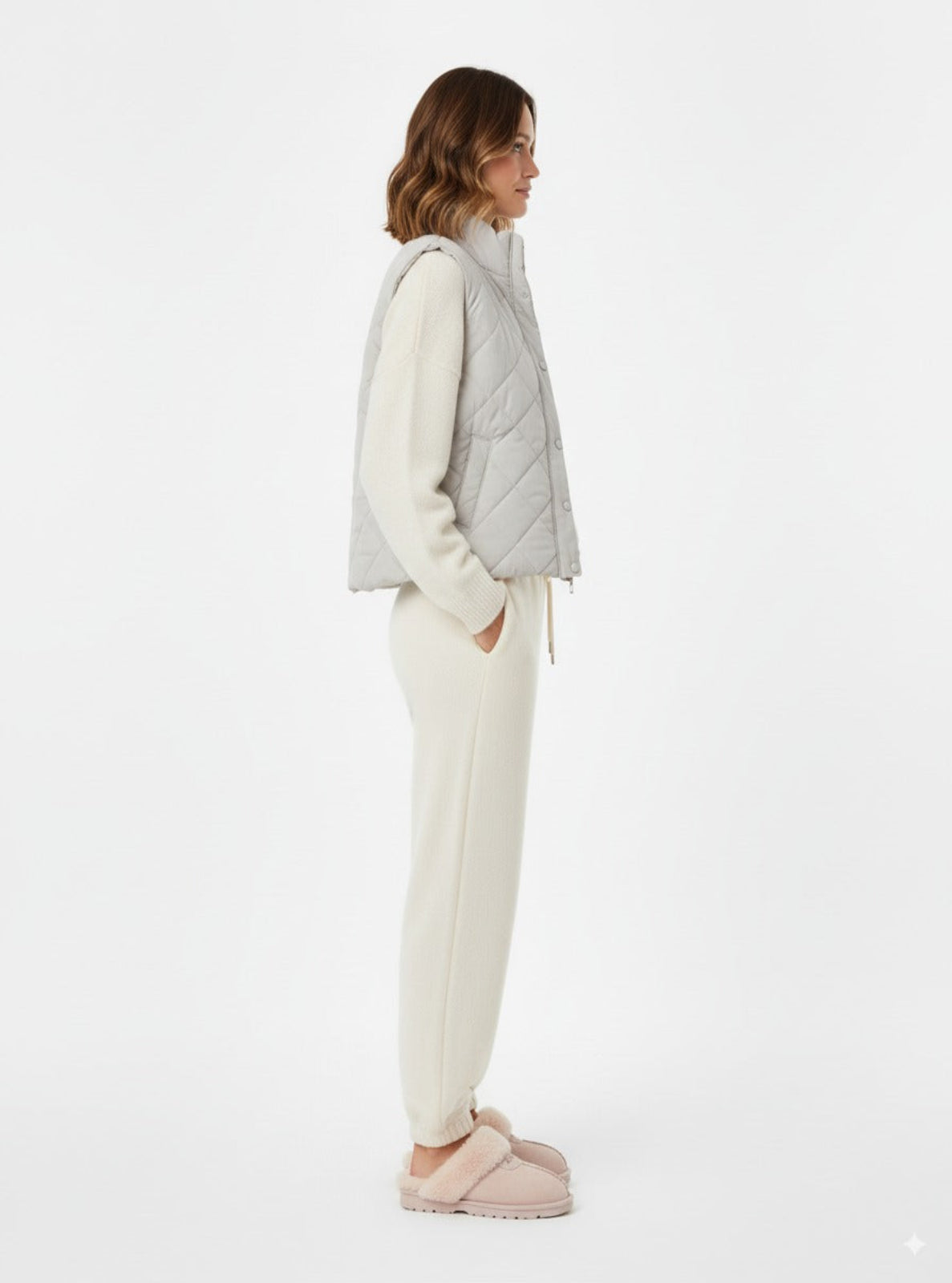 Woman wearing a light gray quilted vest over a white sweater and cream pants on a white background