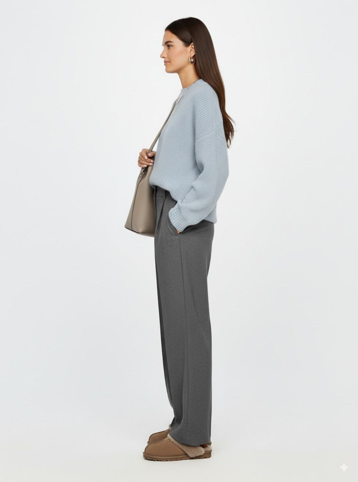 Woman wearing a light blue sweater and gray pants, holding a beige handbag on a white background