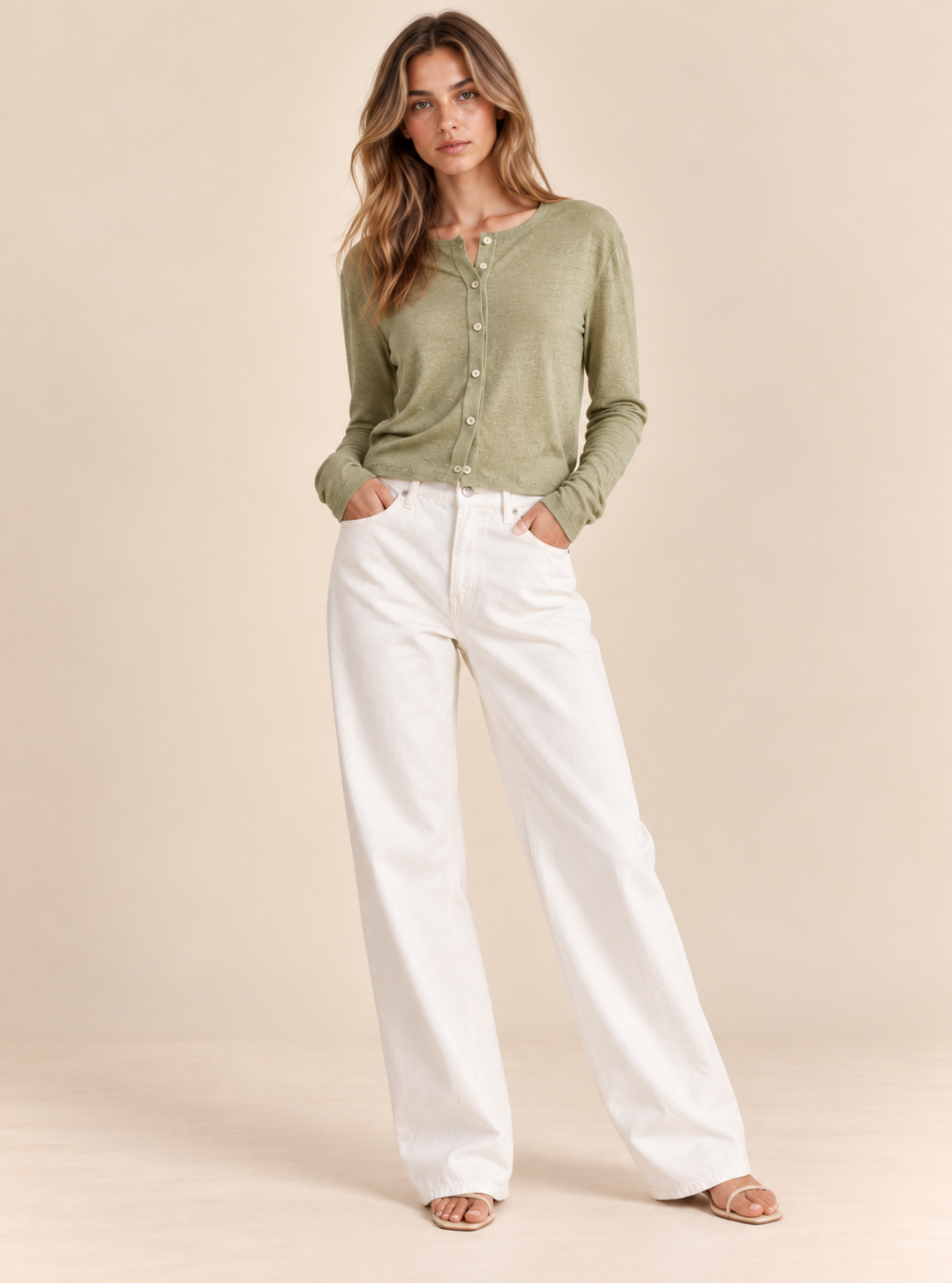 A woman modeling a sage green long-sleeve henley cardigan and white wide-leg pants against a solid beige studio background.