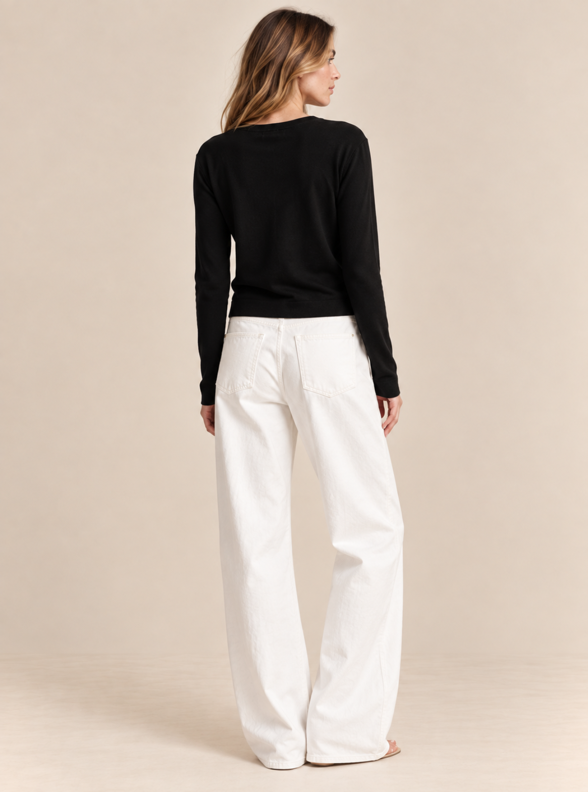 A woman shown from the back wearing a black long-sleeve top and white wide-leg pants, standing against a plain beige studio background.