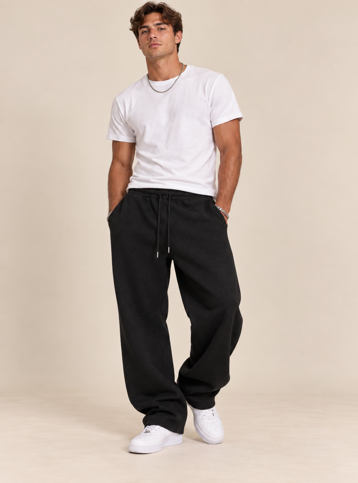 A man modeling a pair of black sweatpants with a white t-shirt and white sneakers, standing against a solid beige studio background.