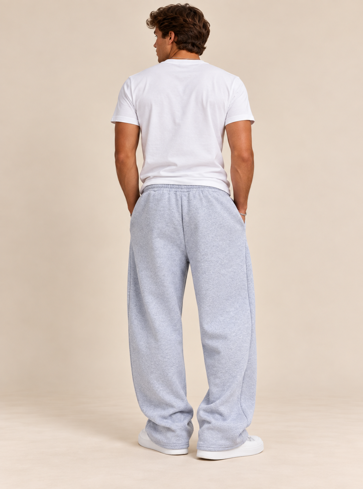 A man viewed from behind wearing a white t-shirt and light heather gray sweatpants, standing against a solid beige studio background.