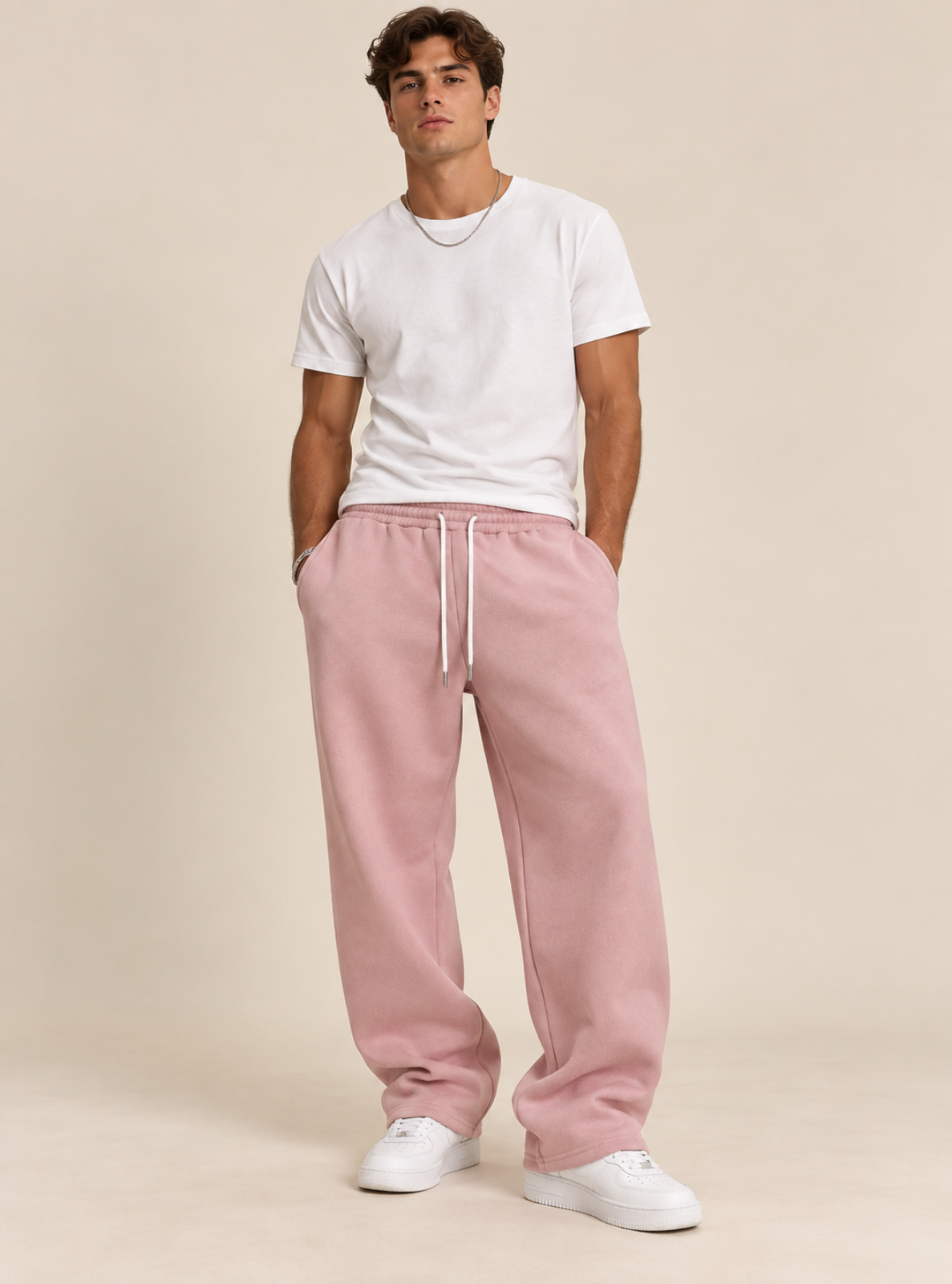A man modeling a pair of loose-fit dusty pink sweatpants with a white drawstring, paired with a white t-shirt and white sneakers, against a solid beige studio background.