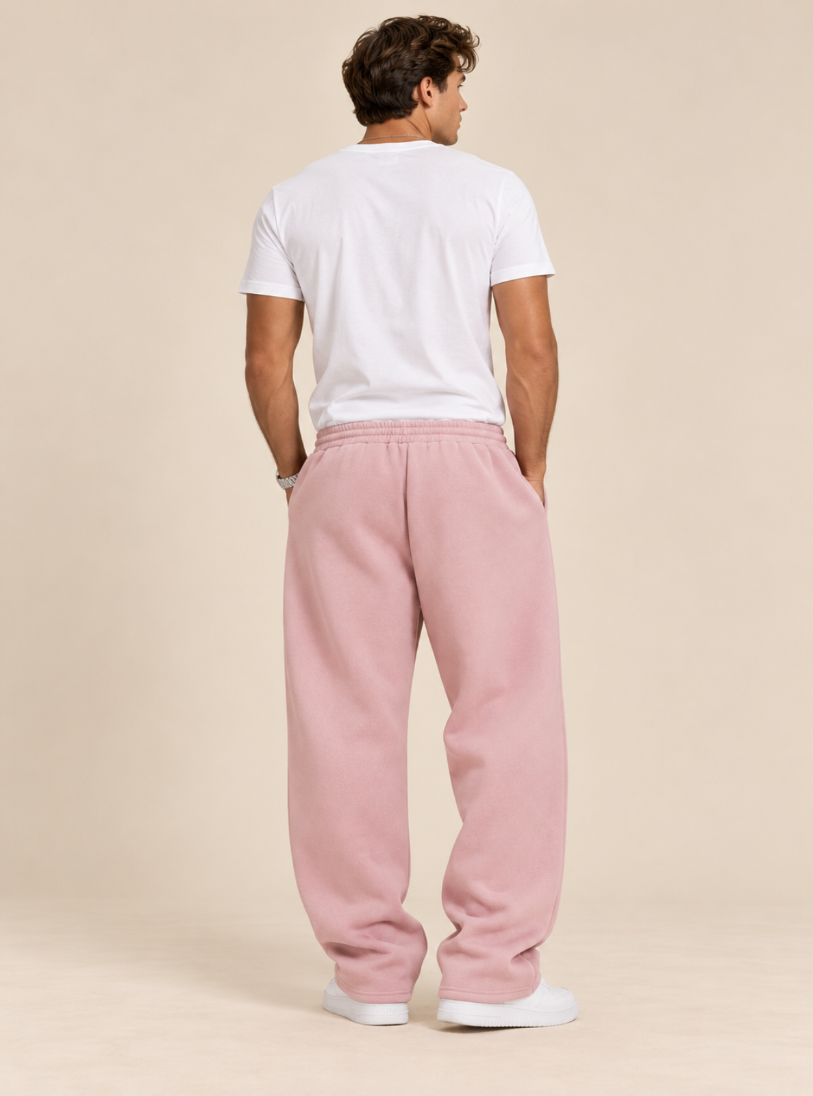 A man viewed from behind wearing a white t-shirt and loose, wide-leg pink sweatpants, standing against a solid beige studio background.