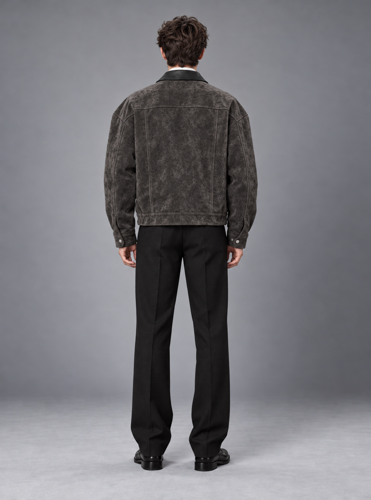 A man viewed from behind wearing a dark textured suede or nubuck jacket over a black shirt and black trousers, standing against a neutral gray studio background.