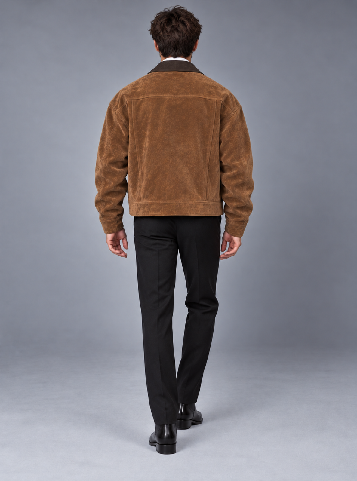 A man viewed from behind wearing a brown suede jacket, black trousers, and black shoes, standing against a neutral gray studio background.