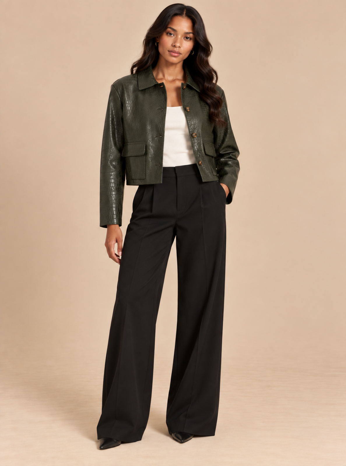 A woman modeling a dark olive green faux-leather cropped jacket over a white top and black wide-leg trousers, standing against a solid beige studio background.