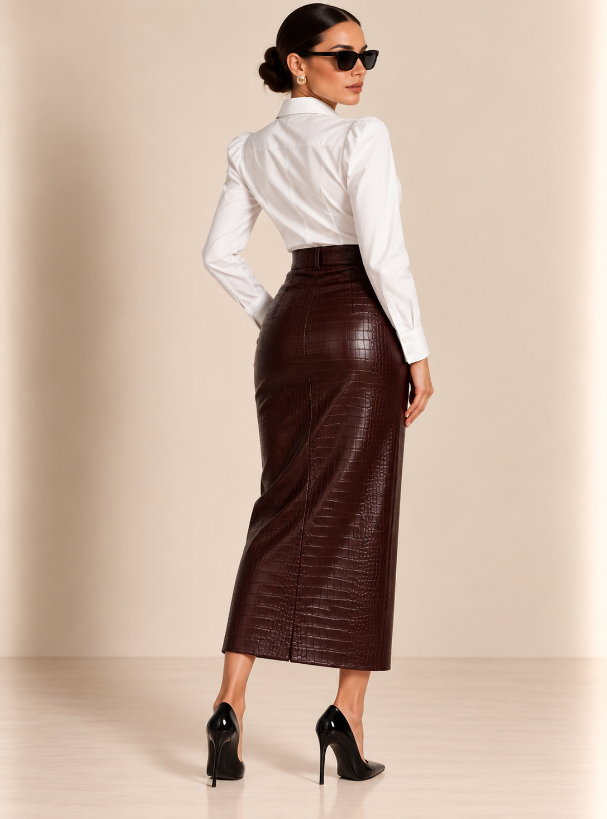 A woman photographed from the back wearing a white long-sleeve blouse, a fitted dark brown faux-leather midi skirt with a crocodile-embossed texture, black sunglasses, and black high heels against a warm beige studio backdrop.