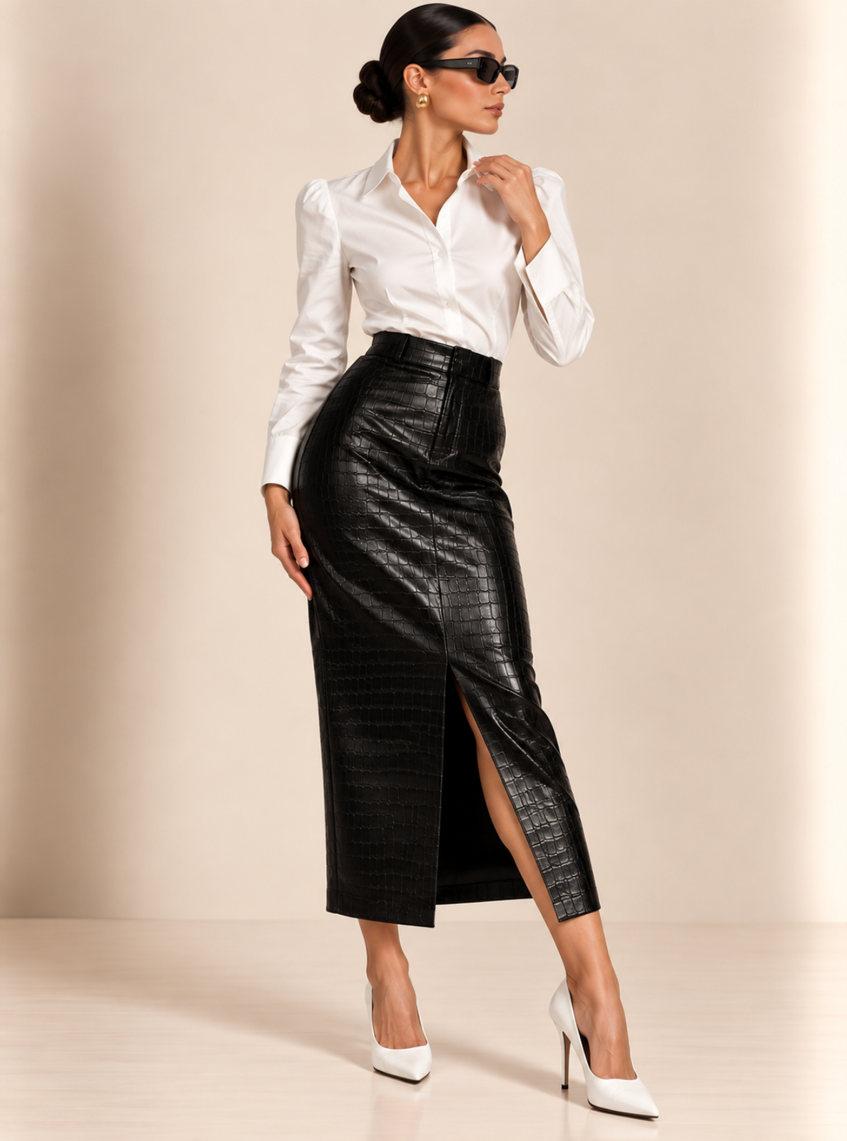 A woman models a high-waisted black faux-leather midi skirt with a front slit, paired with a white button-down shirt and white pointed-toe heels, against a warm beige studio background.