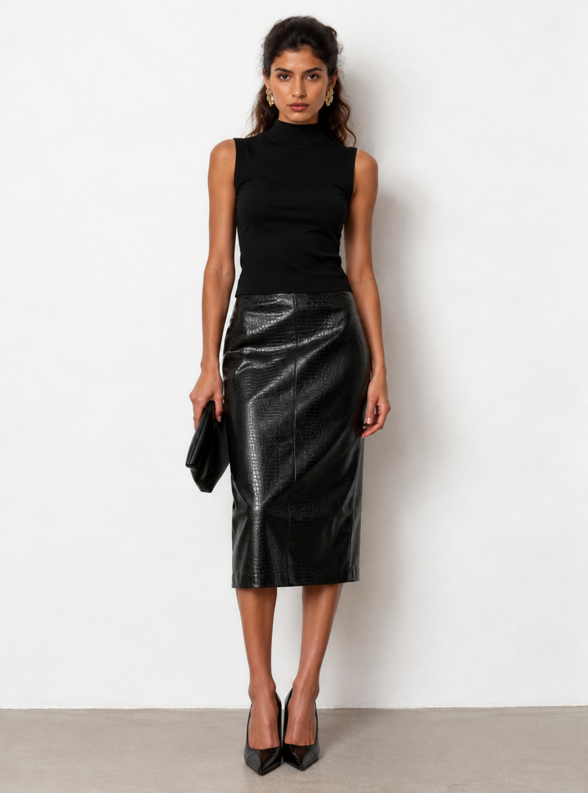 A woman stands against a plain white wall wearing a black sleeveless mock-neck top and a black faux-leather knee-length skirt, holding a black clutch and wearing black pointed heels.
