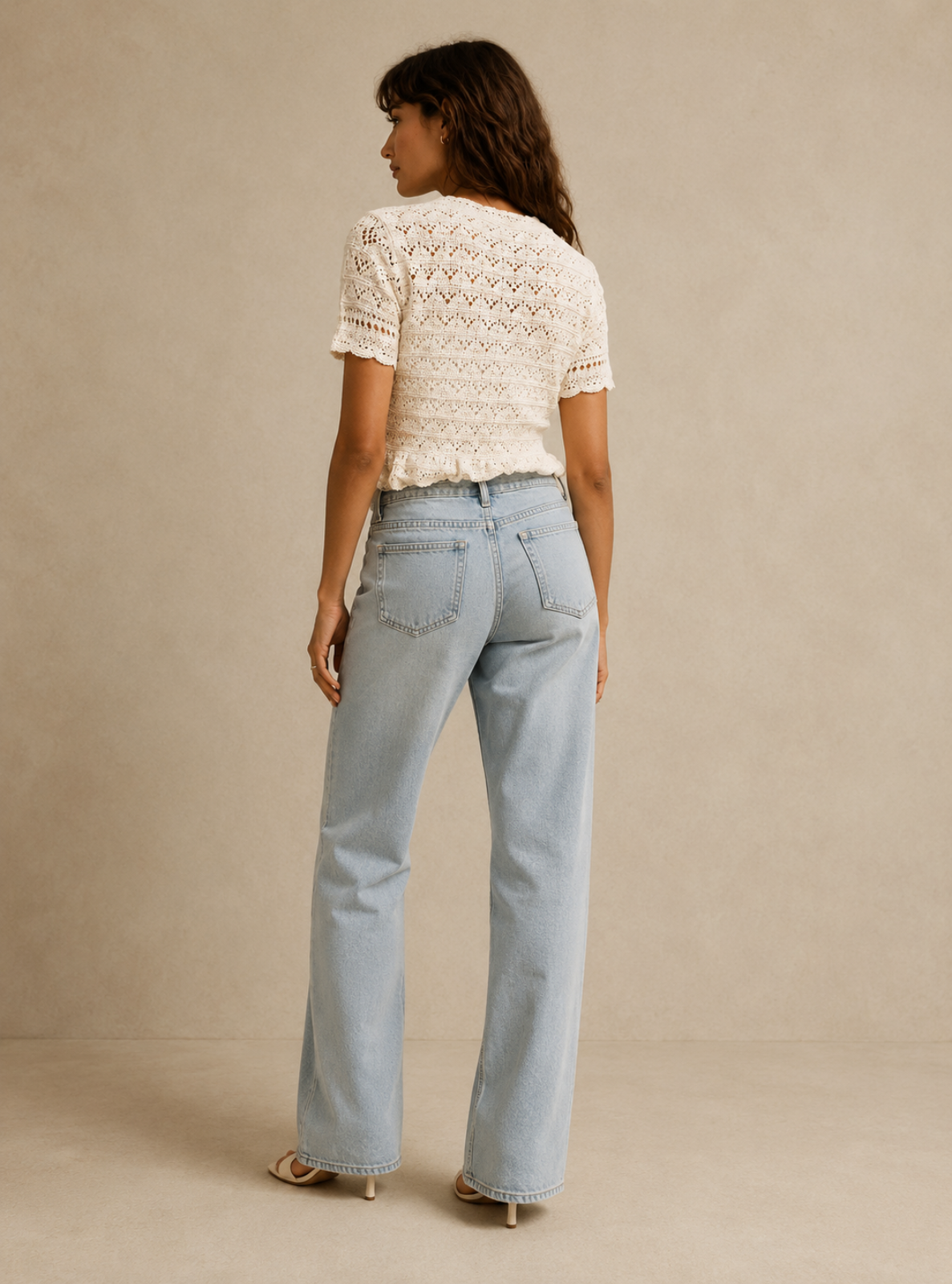 A woman shown from the back wearing a cream crochet short-sleeve top and light-wash blue straight-leg jeans, standing against a neutral beige studio backdrop.