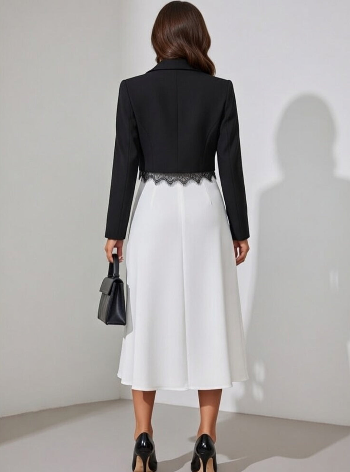 Woman wearing a black blazer and white skirt holding a black handbag against a plain background