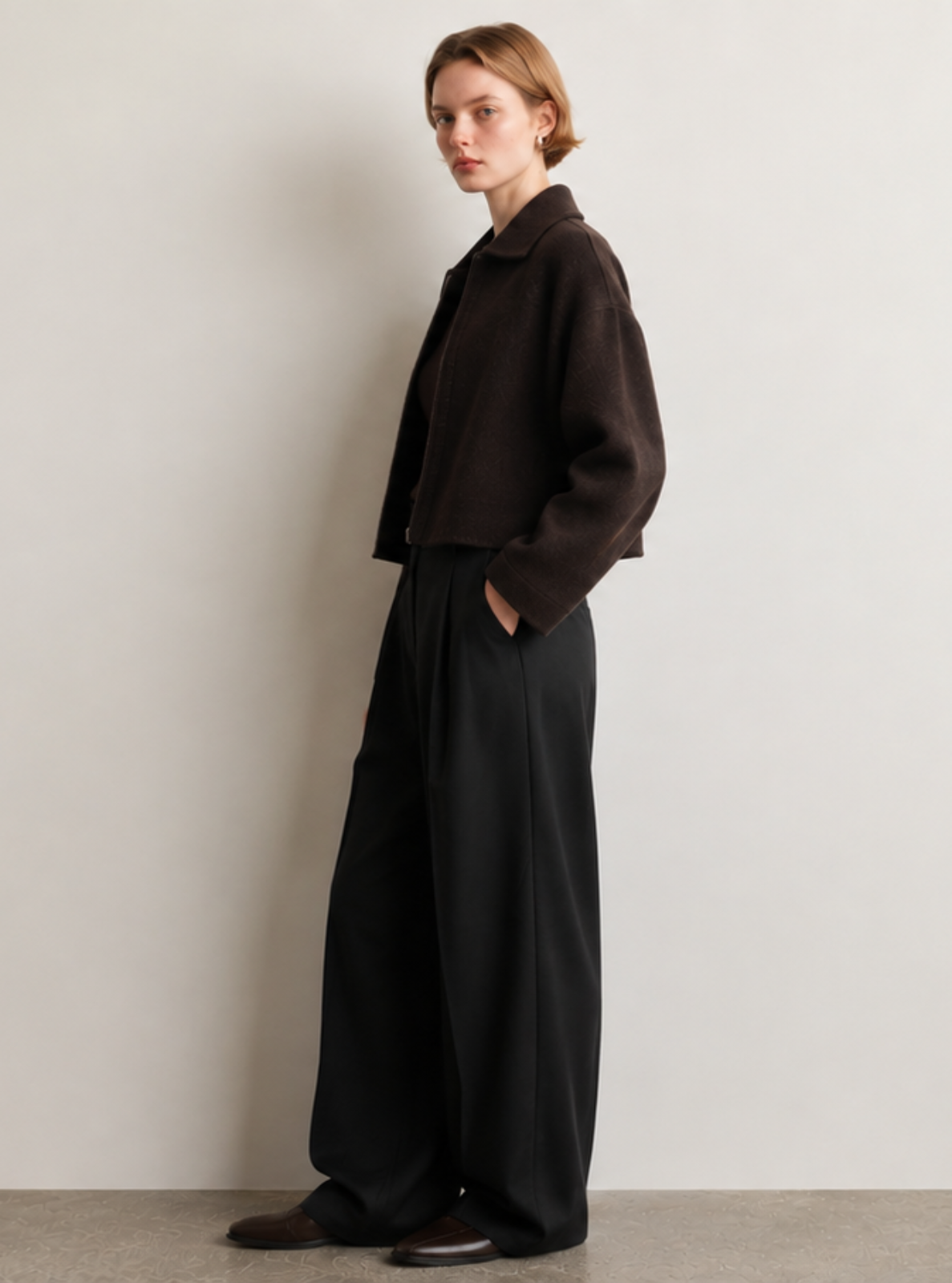 A model stands in profile wearing a dark brown cropped jacket and wide-leg black trousers against a minimalist off-white wall and concrete floor.