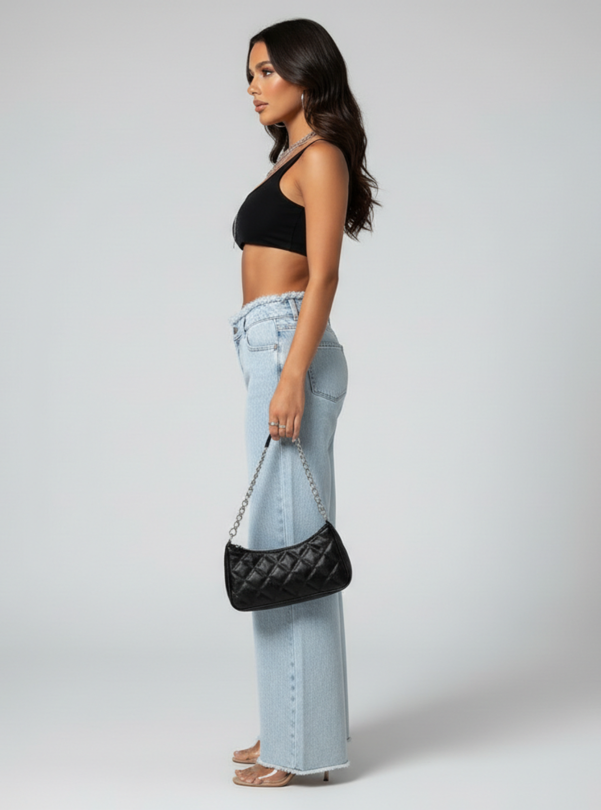 Woman wearing light blue jeans and a black top, holding a black handbag on a white background