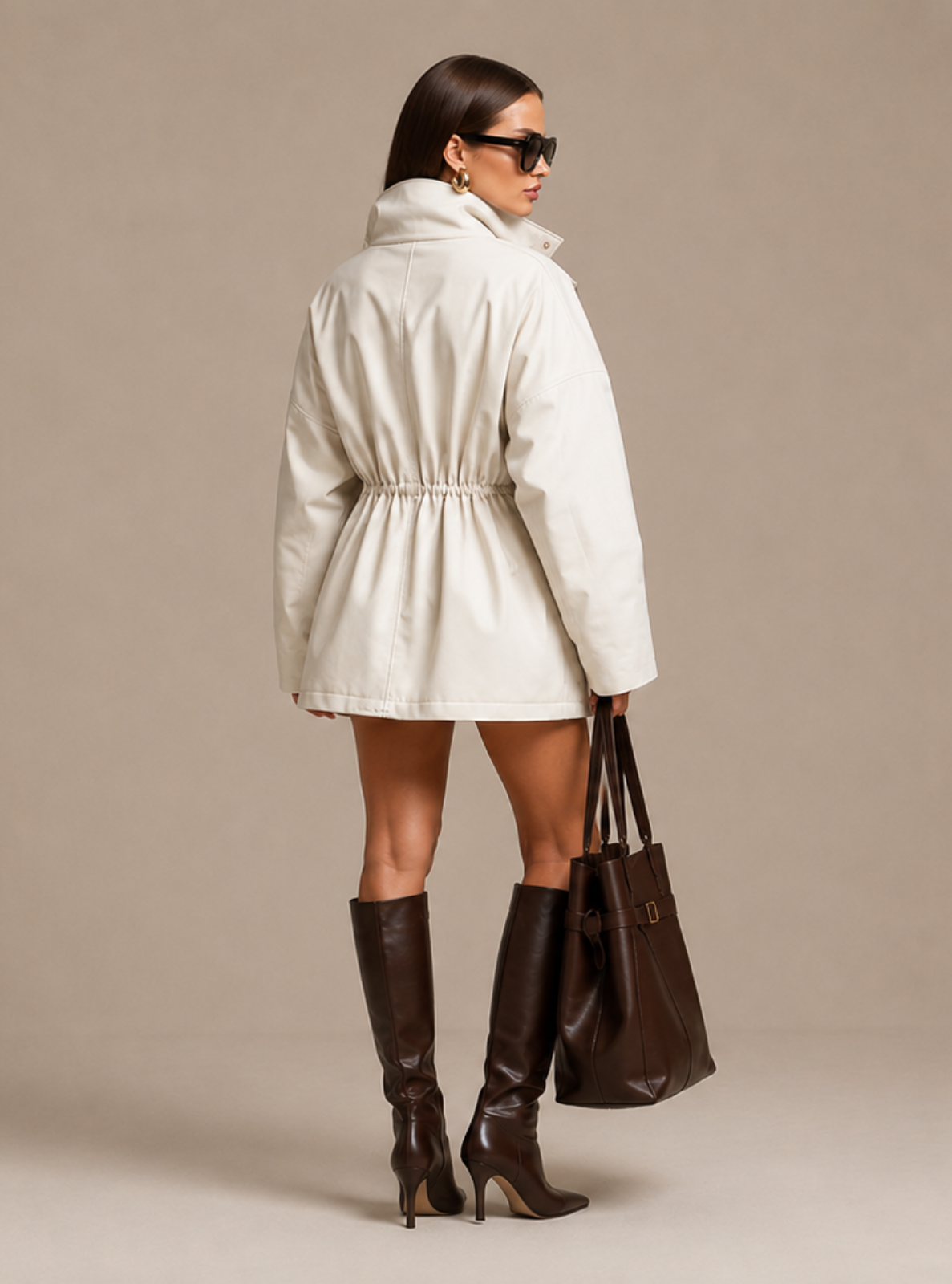 A woman photographed from the back wearing a cream-colored hooded jacket with a drawstring waist, paired with dark brown knee-high boots and carrying a dark brown tote bag, set against a solid beige studio background.
