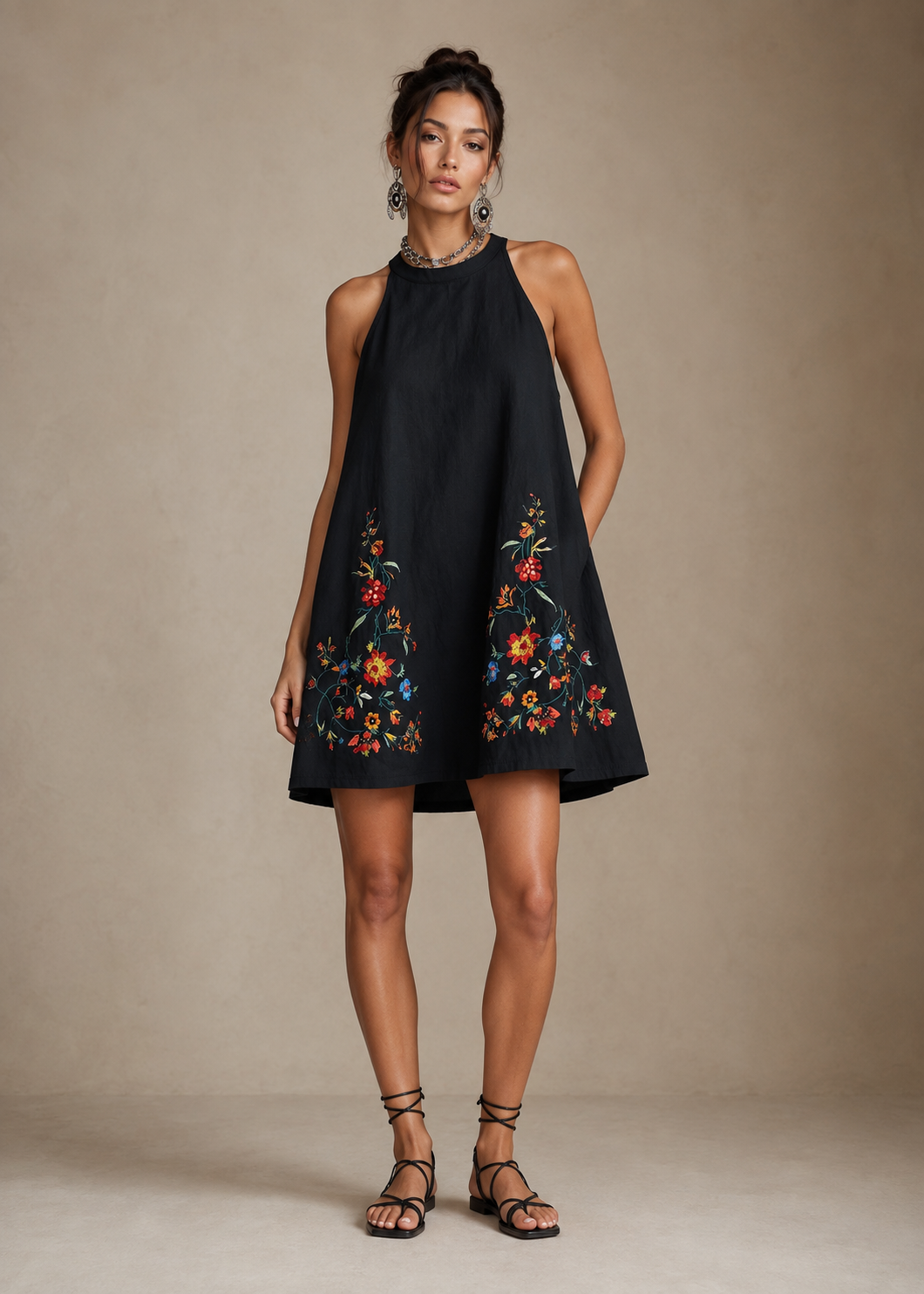 A woman modeling a sleeveless black halter mini dress with colorful floral embroidery near the hem, paired with black strappy sandals, against a neutral beige studio backdrop.