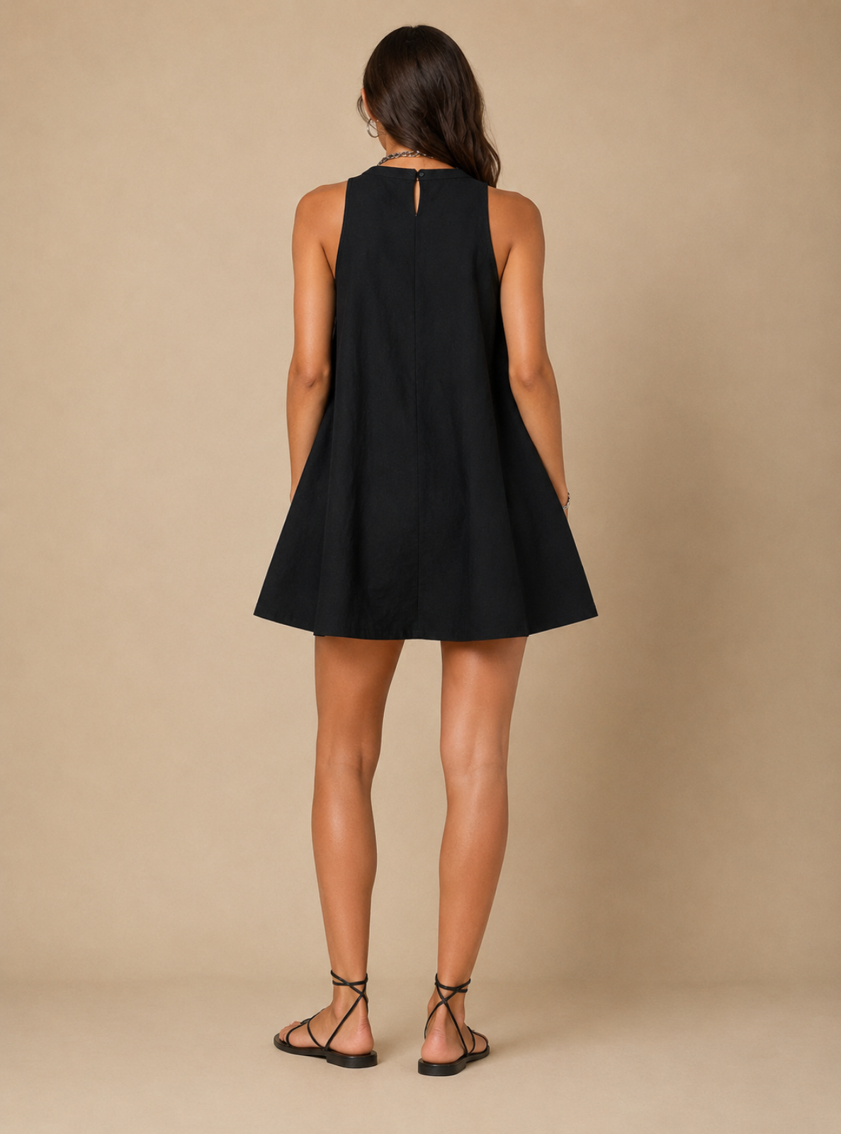 A woman shown from the back wearing a sleeveless black mini dress with a flared skirt and a small keyhole cutout at the back, paired with black strappy flat sandals against a plain beige studio backdrop.