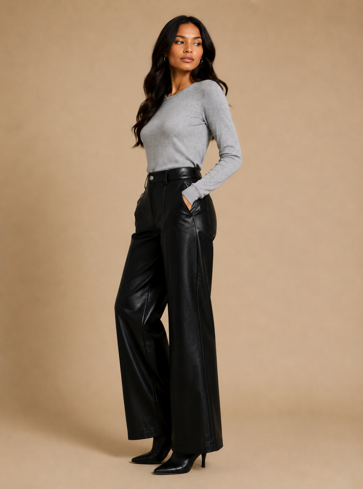A woman modeling black wide-leg faux leather pants with a black belt, paired with a light gray long-sleeve top and black heels, against a solid beige studio background.