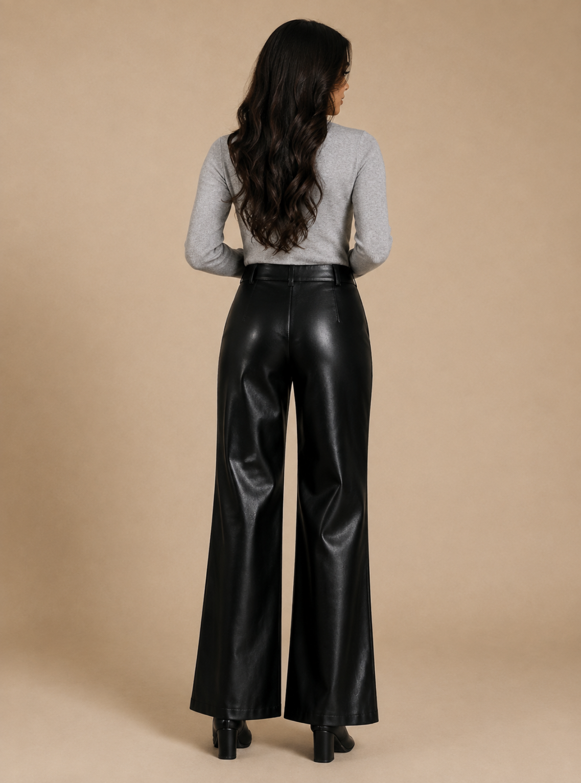 A woman viewed from behind wearing high-waisted black wide-leg faux leather pants and a light gray long-sleeve top, standing against a solid beige studio background.