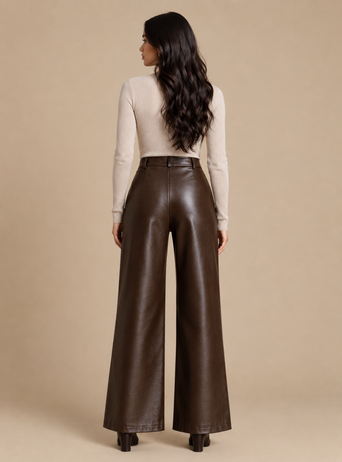 A woman shown from the back wearing high-waisted, wide-leg brown faux-leather pants and a cream ribbed long-sleeve top, standing against a solid beige studio background.