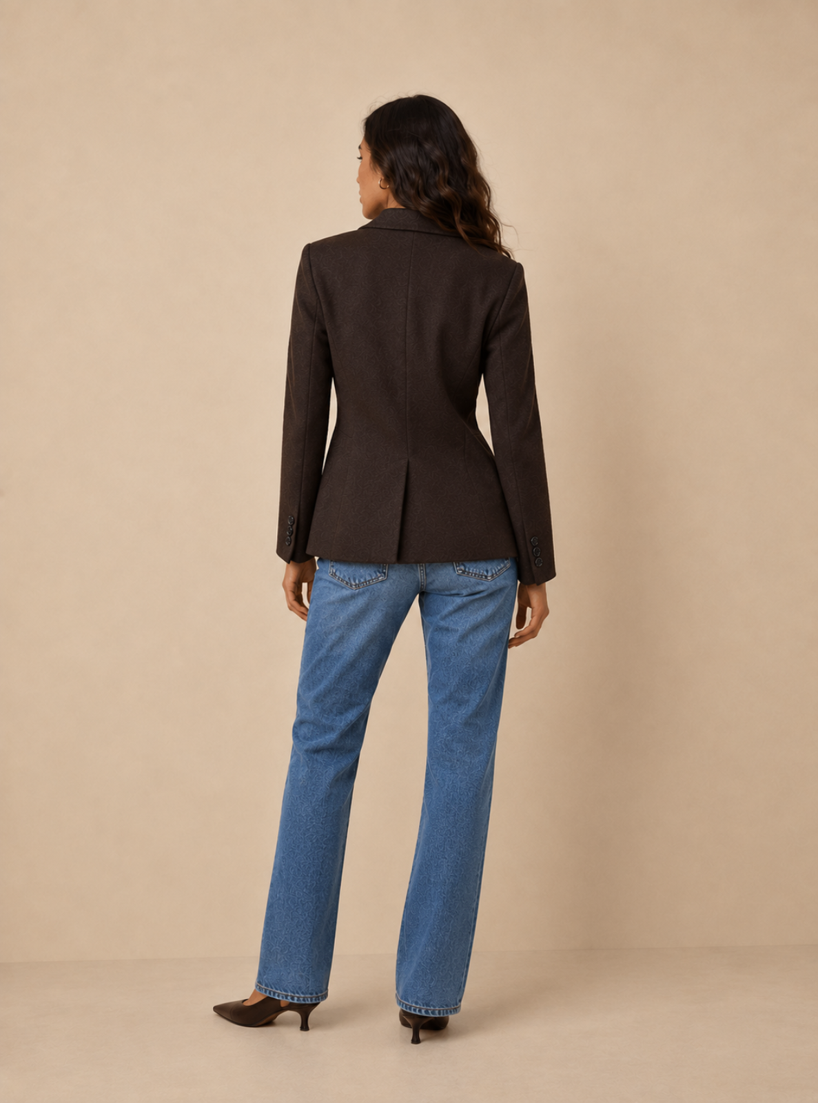 A woman shown from the back wearing a dark brown tailored blazer and blue jeans, standing against a plain beige studio backdrop.