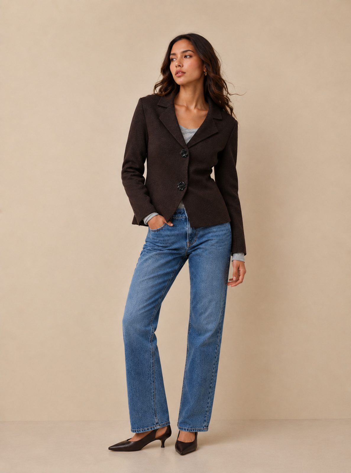 A woman modeling a dark brown tailored blazer with notched lapels and two front buttons, paired with blue straight-leg jeans and black pointed-toe heels, against a plain beige studio backdrop.